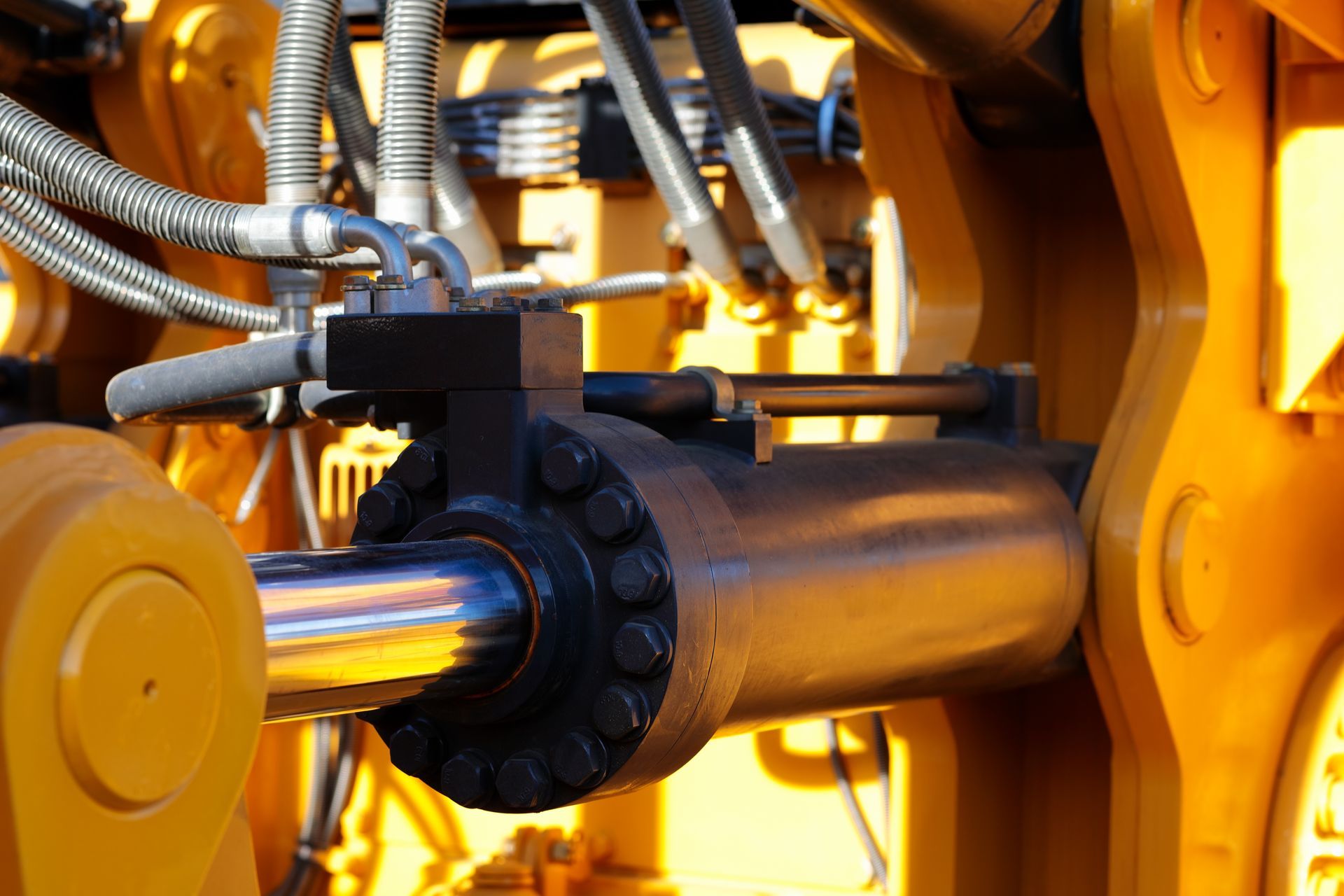 A close-up of a yellow industrial hydraulic cylinder attached to machinery with metal hoses and a silver piston rod.