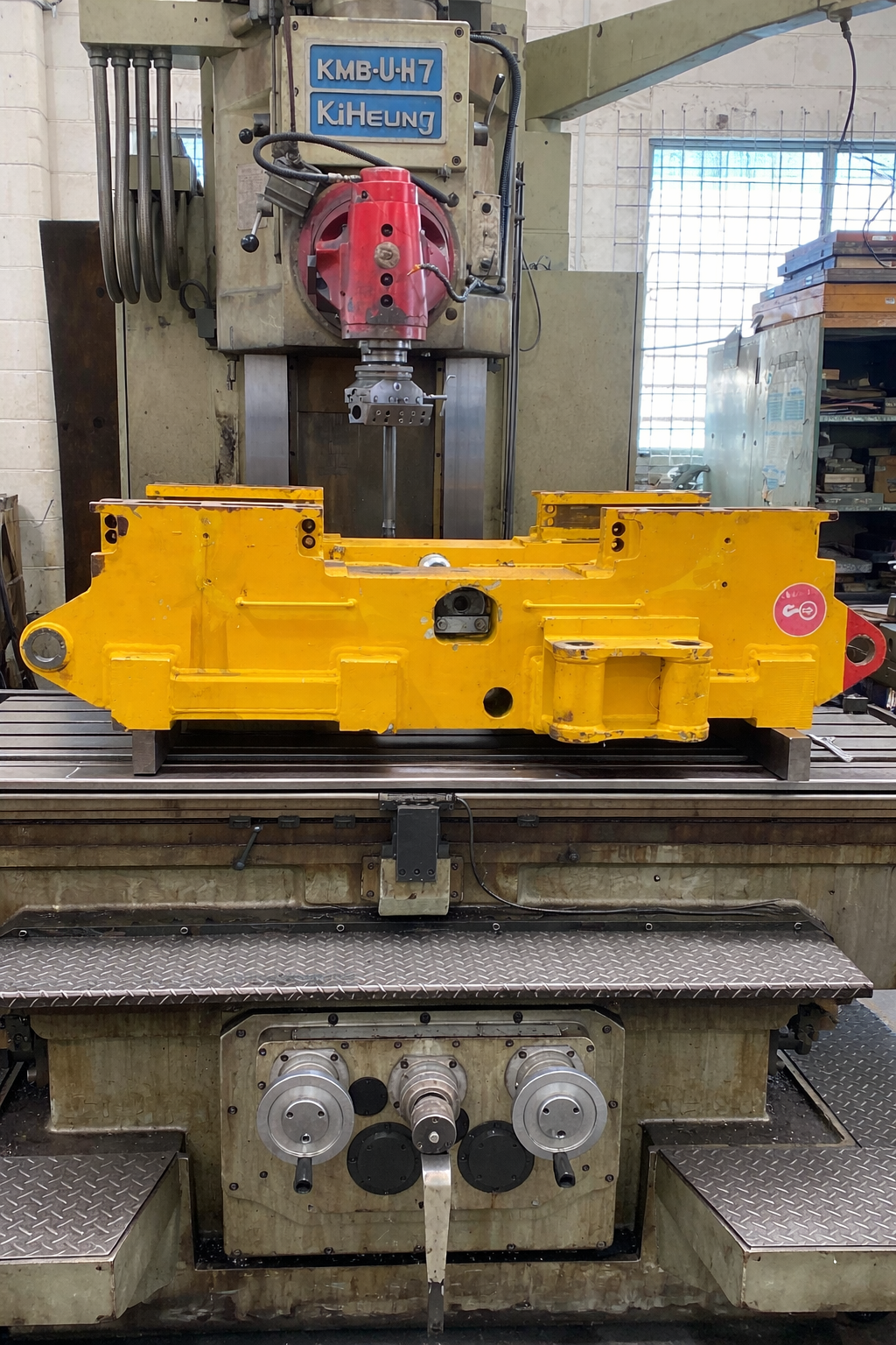 Yellow industrial machine part on a metal worktable inside a workshop, with a drill press behind it. Yellow industrial machine part on a metal worktable inside a workshop, with a drill press behind it.