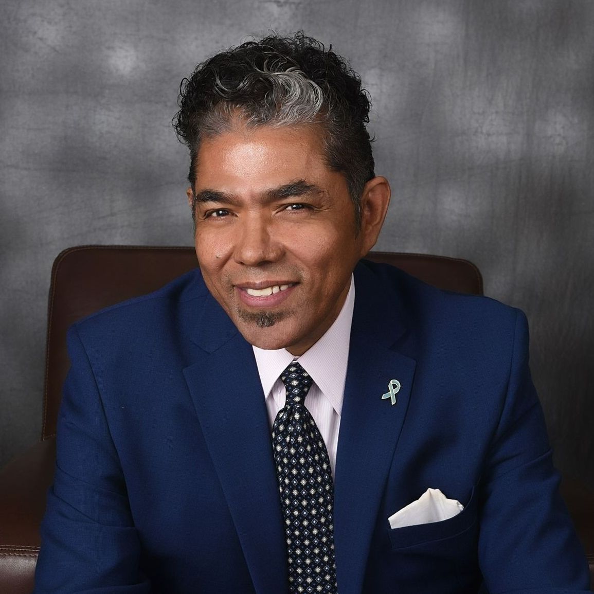Man in blue suit, smiling. Wearing tie, pocket square, and small green ribbon pin.