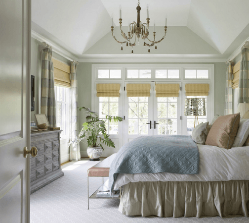 Bedroom with French doors, chandelier, bed with blue quilt, and light-colored walls and curtains.
