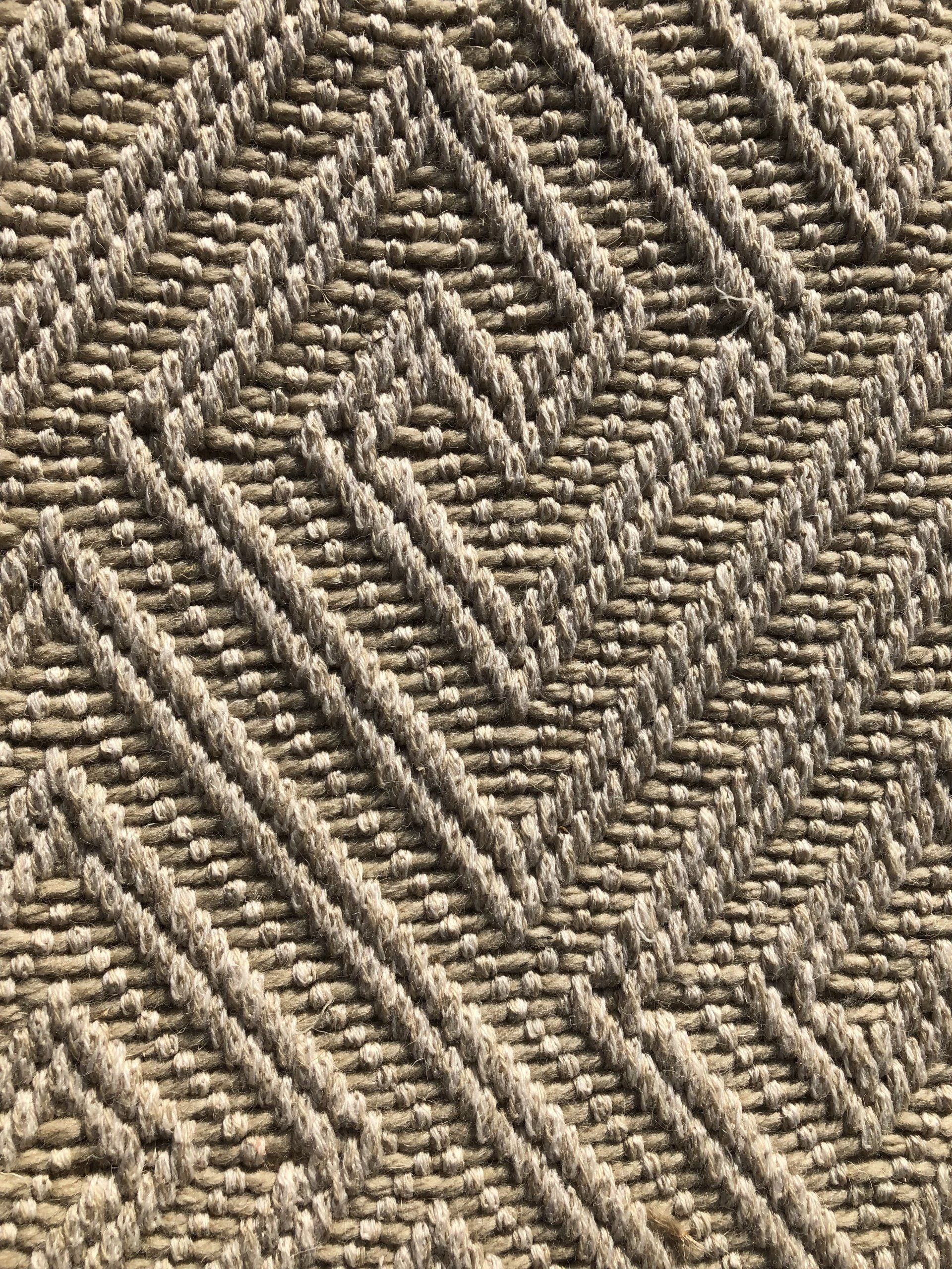 Close-up of a woven rug with a repeating geometric diamond pattern in shades of tan and brown.