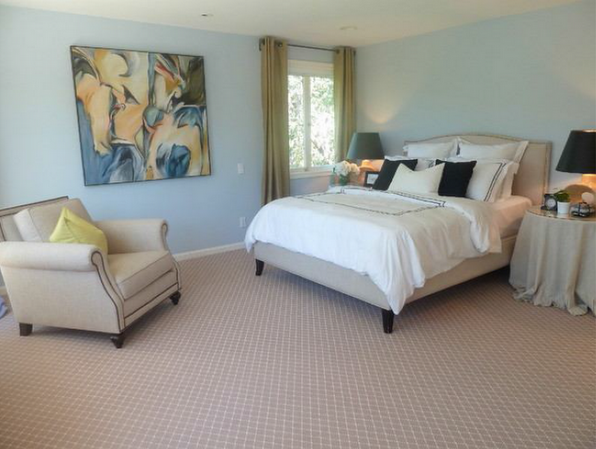 Bedroom with blue walls, beige carpet, bed with white bedding, chair, and artwork.