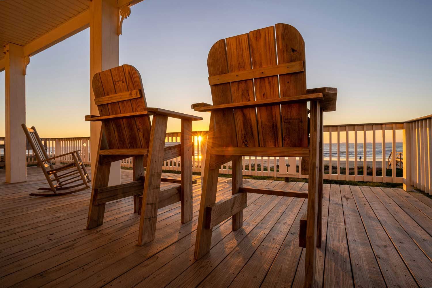 Two wooden chairs are sitting on a wooden deck overlooking the ocean at sunset.