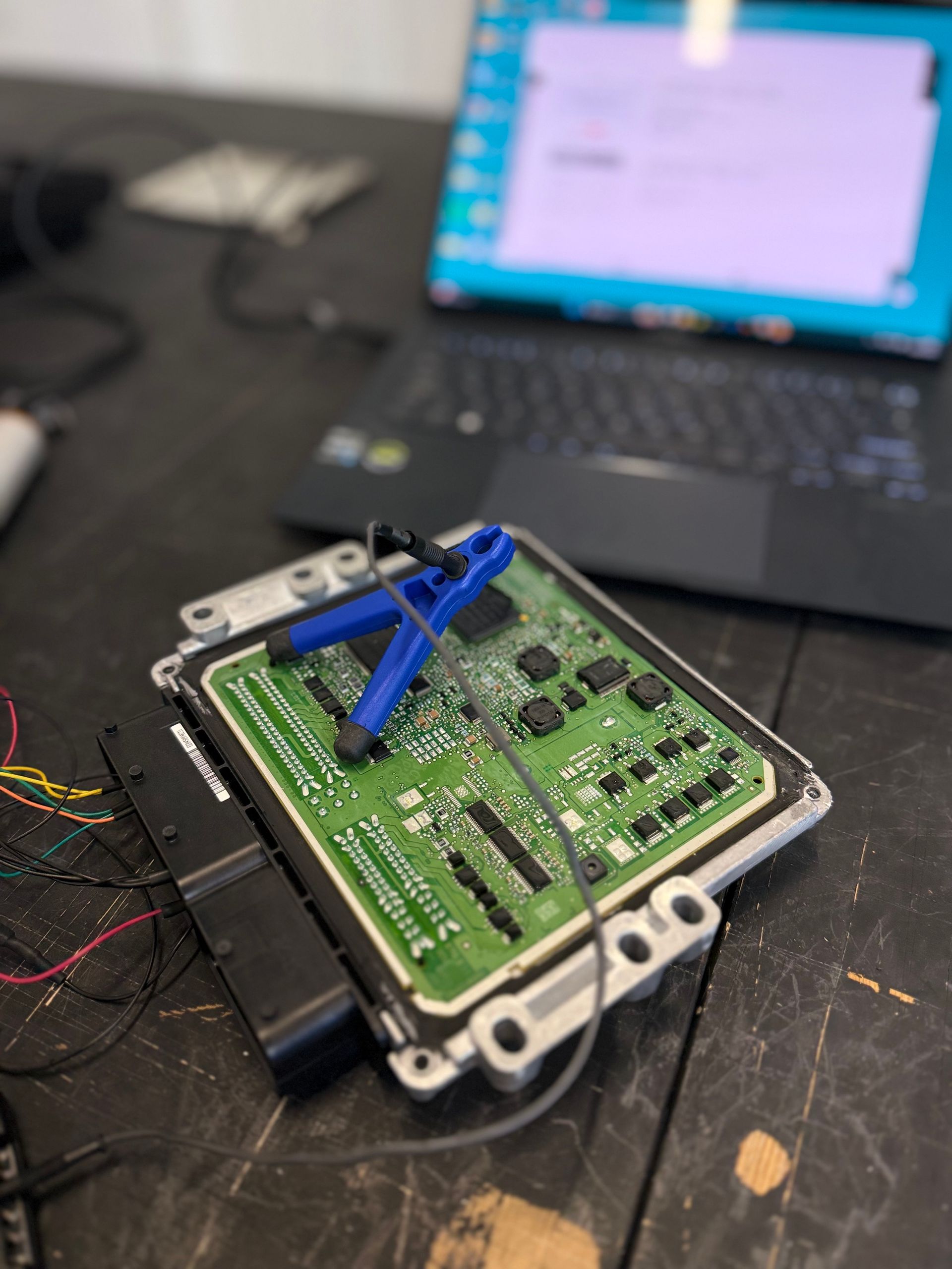 Circuit board with blue clamp and laptop in background.  Wires connected on table.