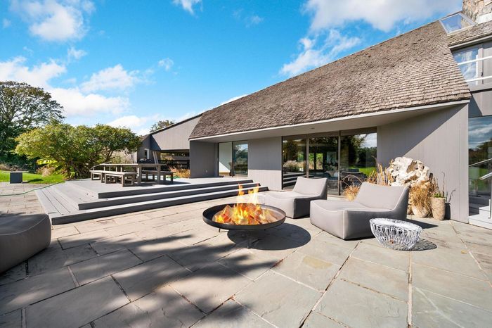 Patio with fire pit, gray chairs, and modern house with large windows under a blue sky.