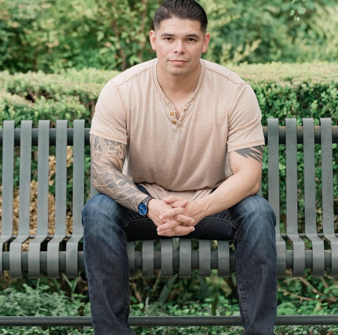 Man with tattoos sits on a bench in a park, hands clasped, looking at the camera.