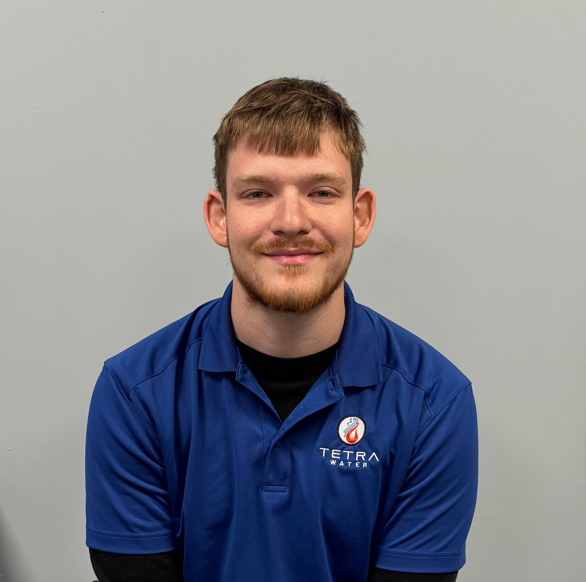 Man with tattoos sits on a bench in a park, hands clasped, looking at the camera.