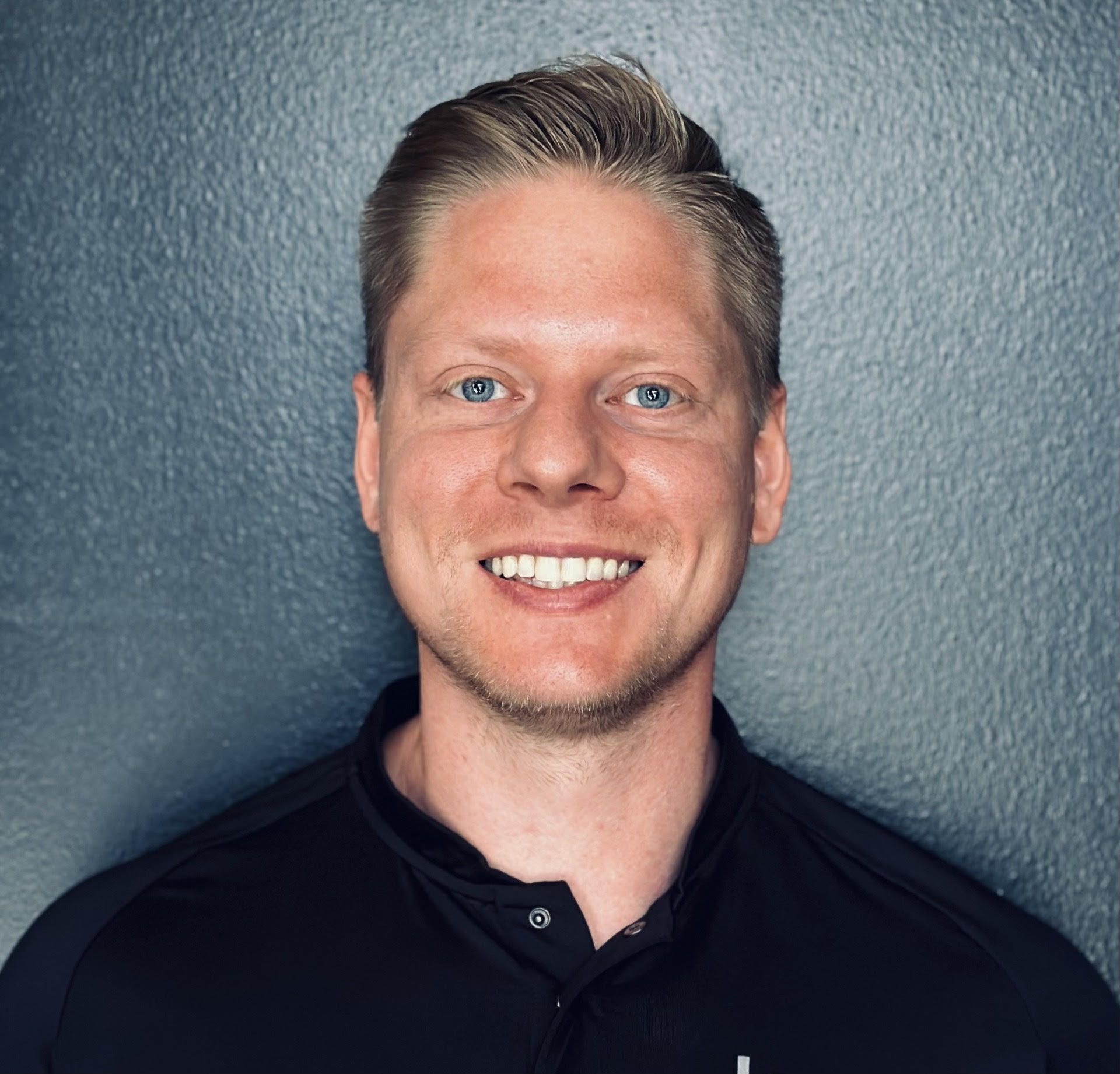 Blond man smiling, head and shoulders shot, black shirt, against a textured gray background.