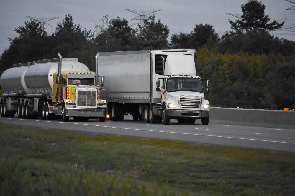 Two semi-trucks on a highway; one yellow tanker truck, one white box truck, overcast sky.