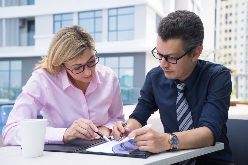 Two people reviewing paperwork outdoors, pointing, with glasses and business attire.