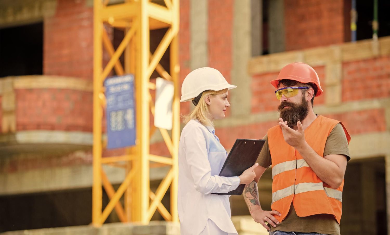 Woman in white hardhat and man in orange hardhat reviewing documents at a construction site. The man is gesturing with his hands.