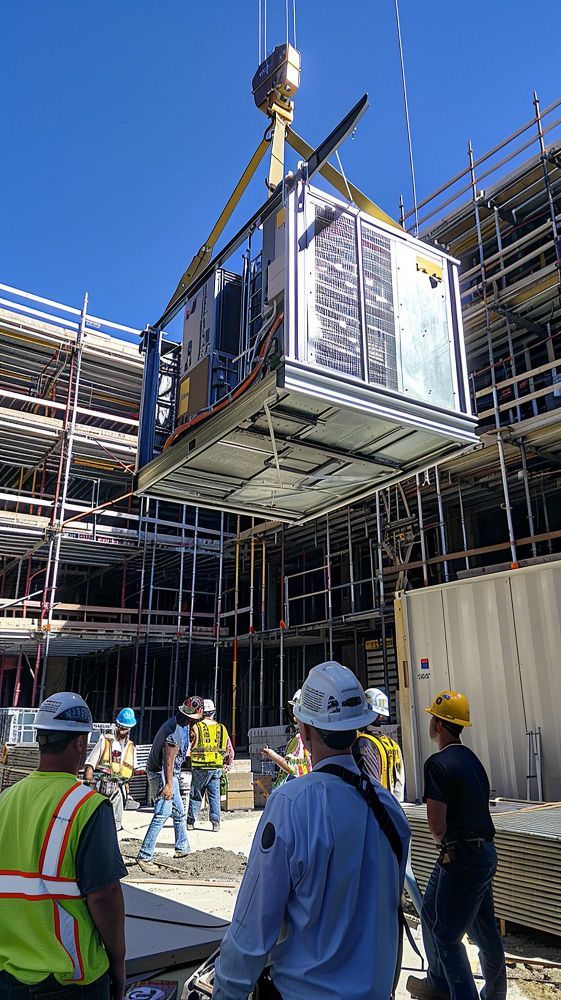 A crane lifts an HVAC unit above construction workers on a building site under a blue sky.