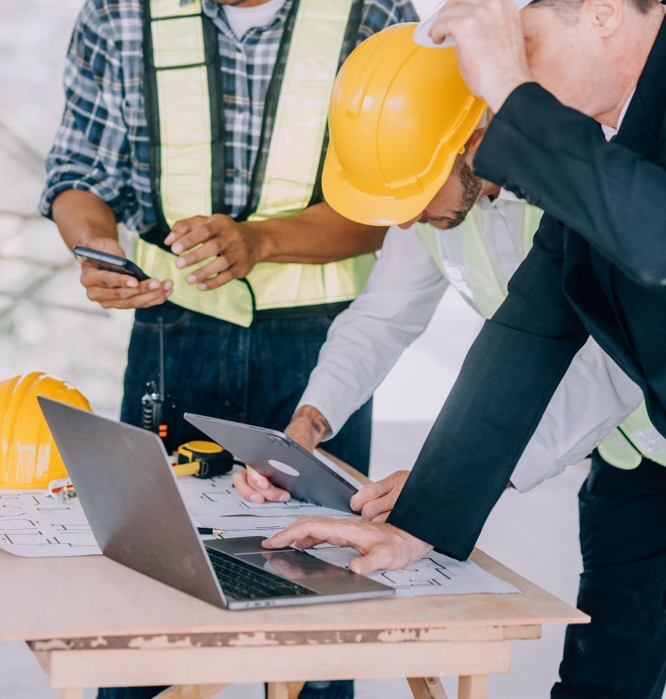 Construction workers review plans at a table, one using a laptop, others holding a tablet and phone; wearing safety vests and helmets.
