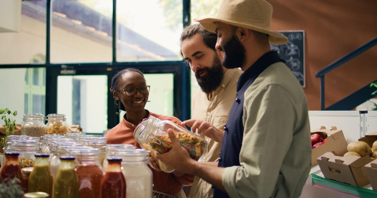 Three people examining a jar of food at a market. The person in a straw hat holds the jar, while the others look on smiling.