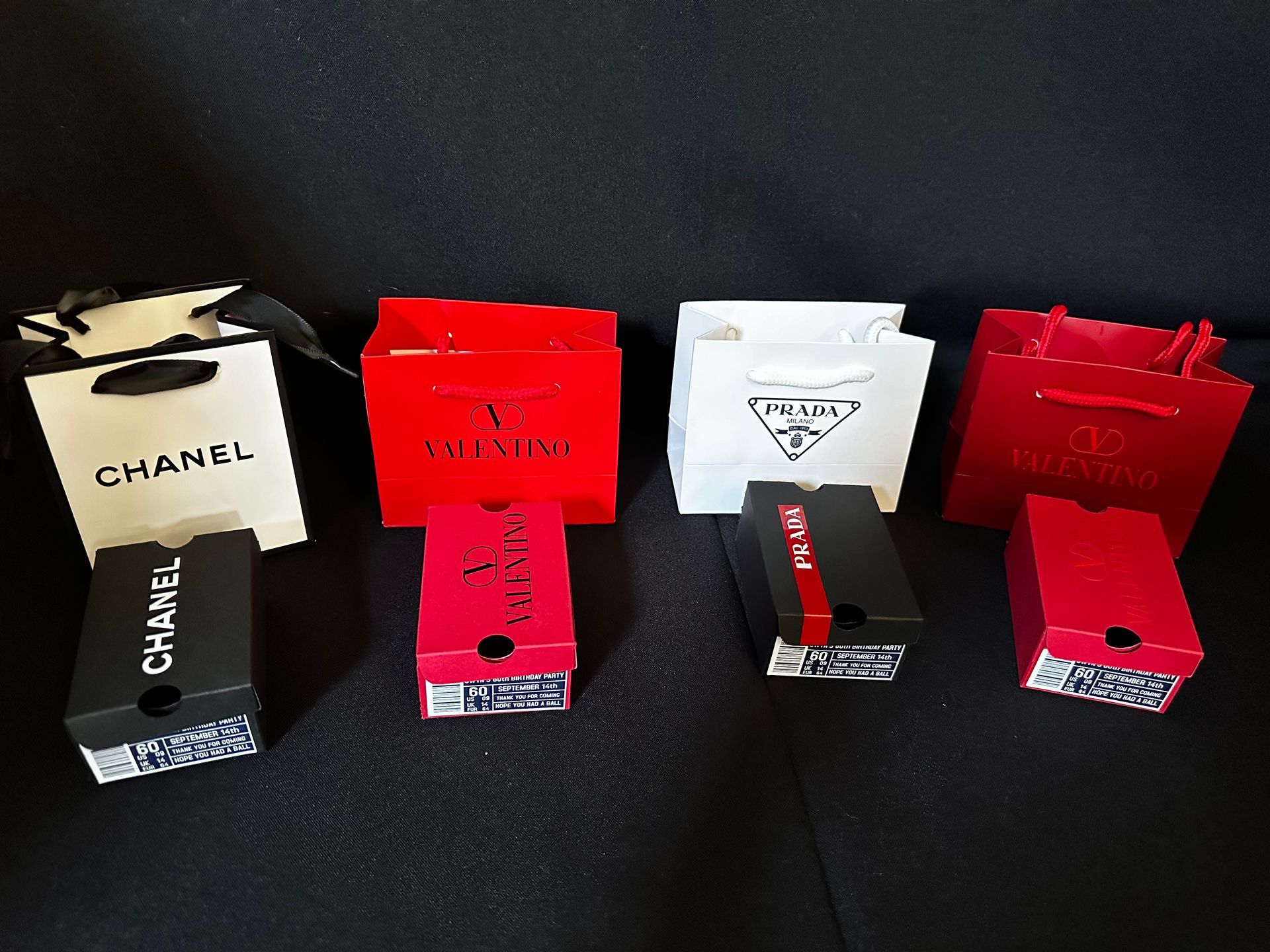 A row of designer brand favor bags and favor shoeboxes are lined up on a table