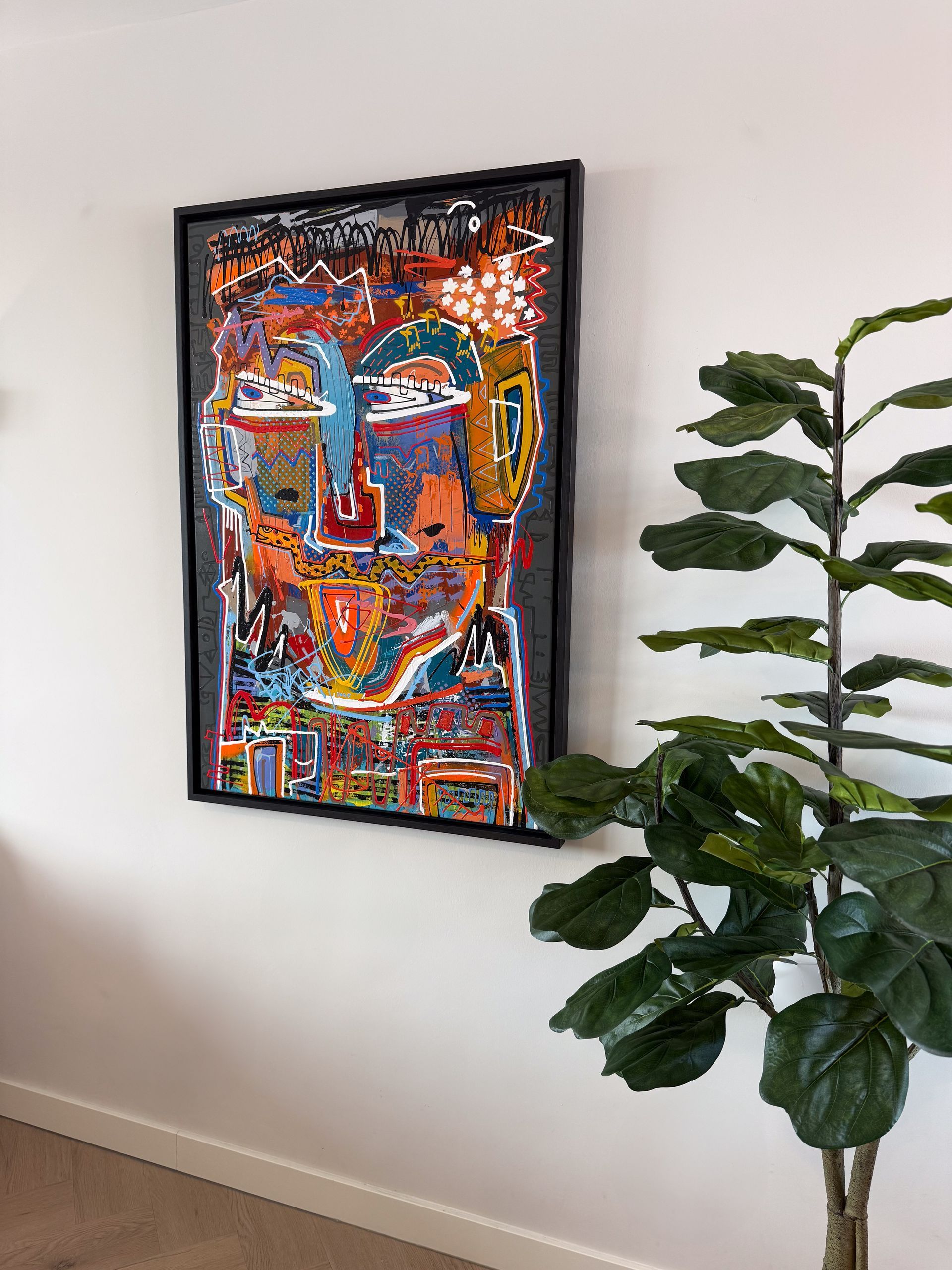 Abstracte kunst in een zwarte lijst op een witte muur; groene plant op de voorgrond.