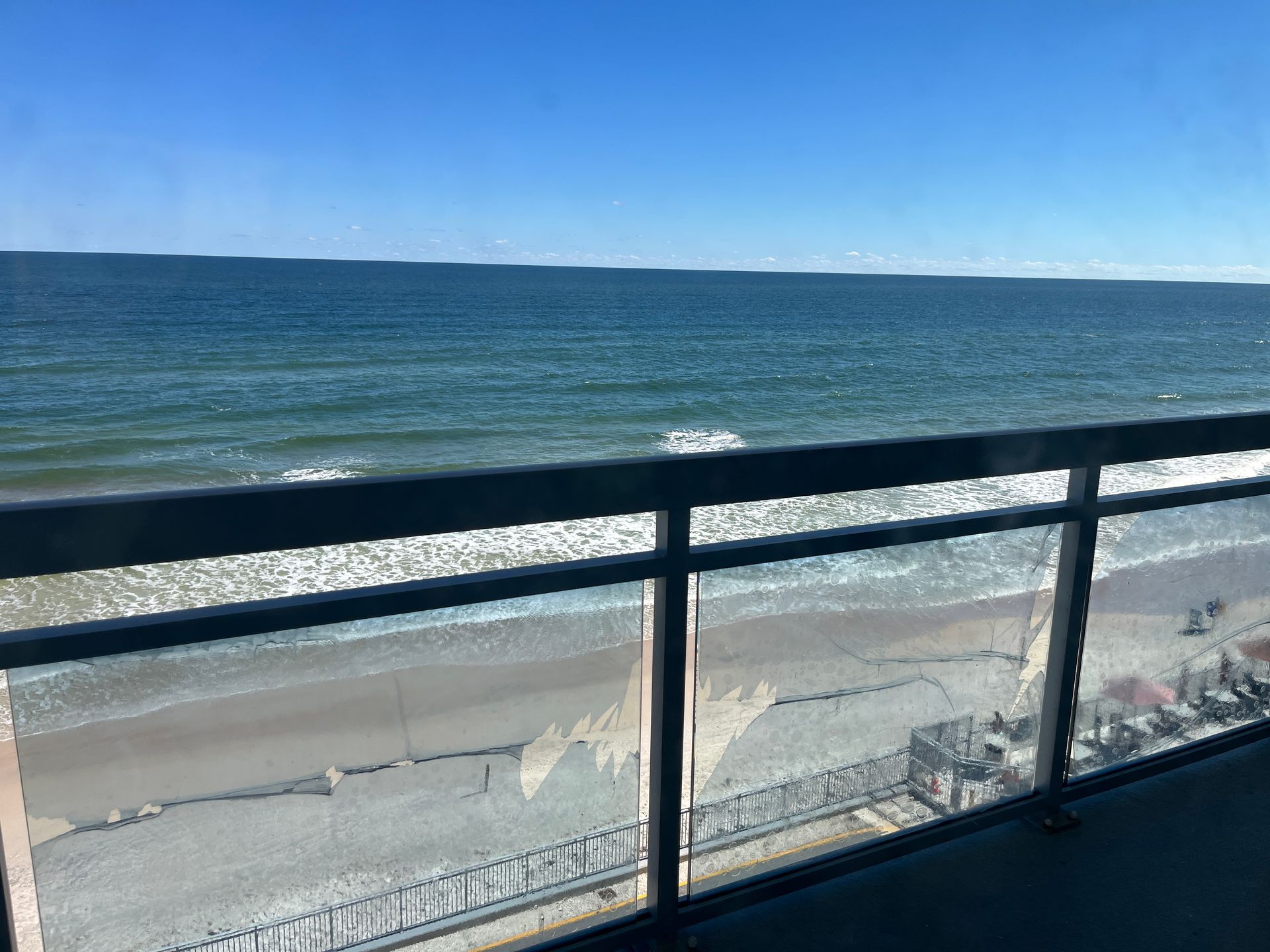 Ocean view from a balcony. Blue sky and water, sandy beach below. Clear railings.