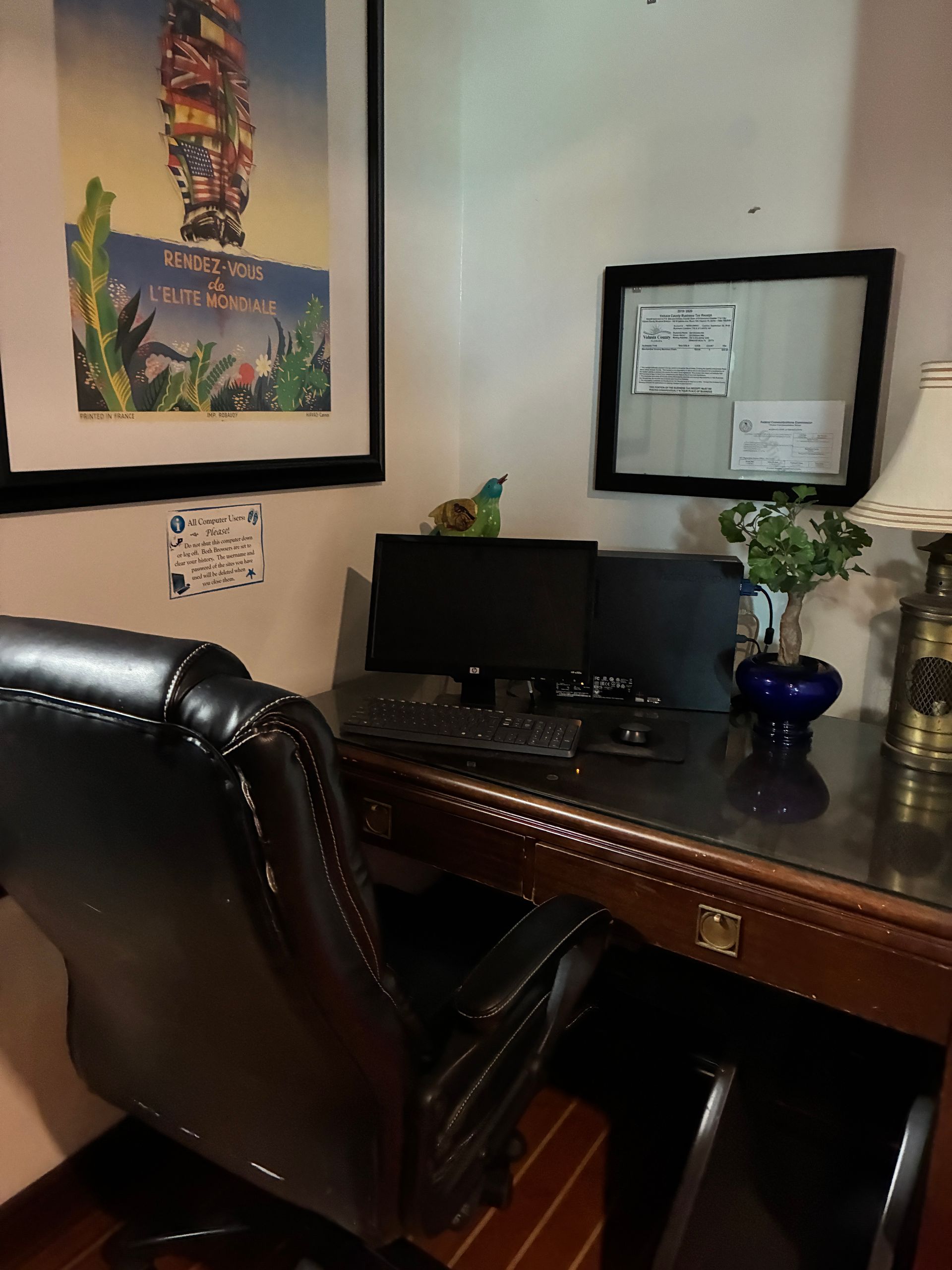 A desk in a corner with a computer, chair, and framed artwork. Dark wood and black leather.