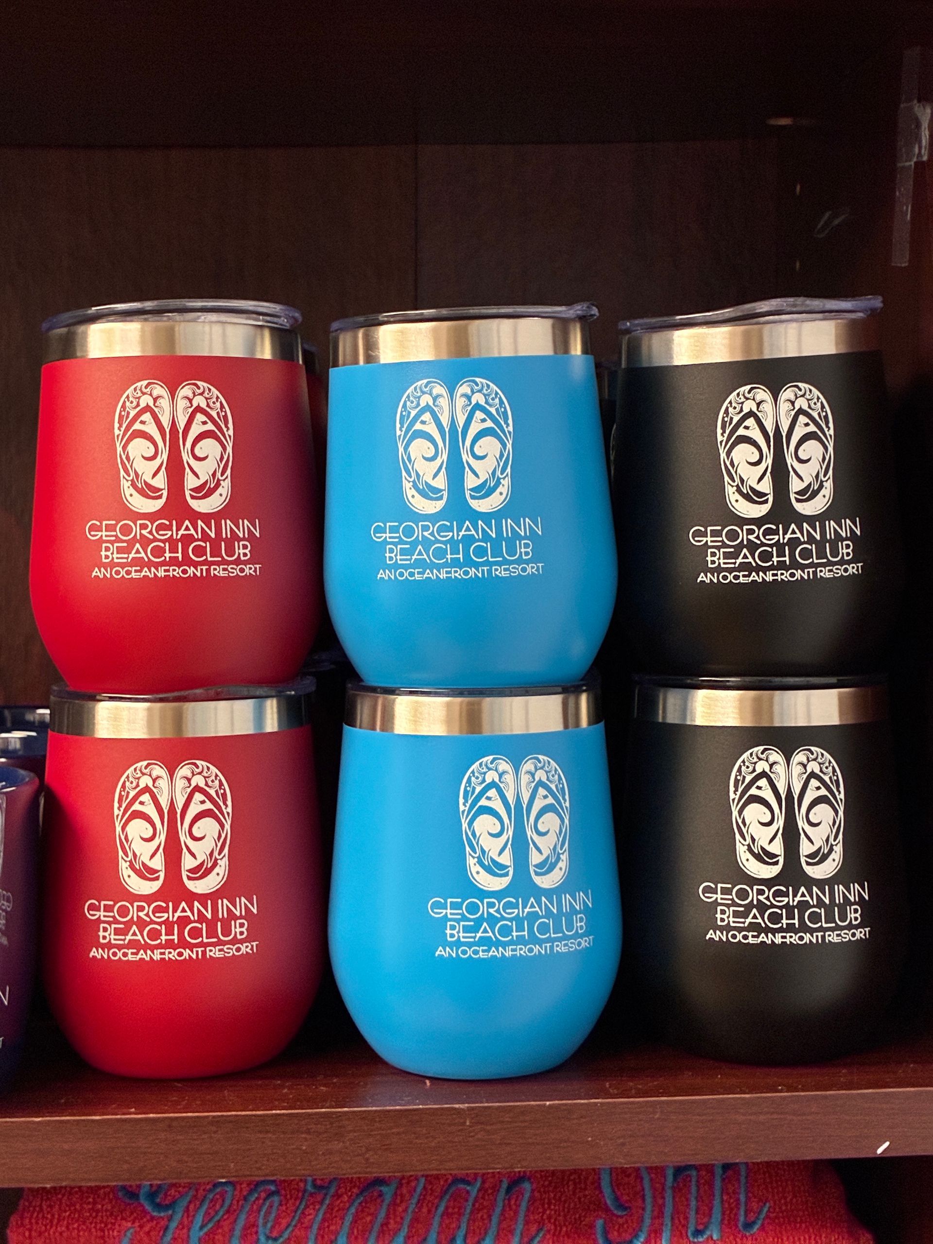 Stemless travel mugs in red, blue, and black with a white logo; on a shelf.