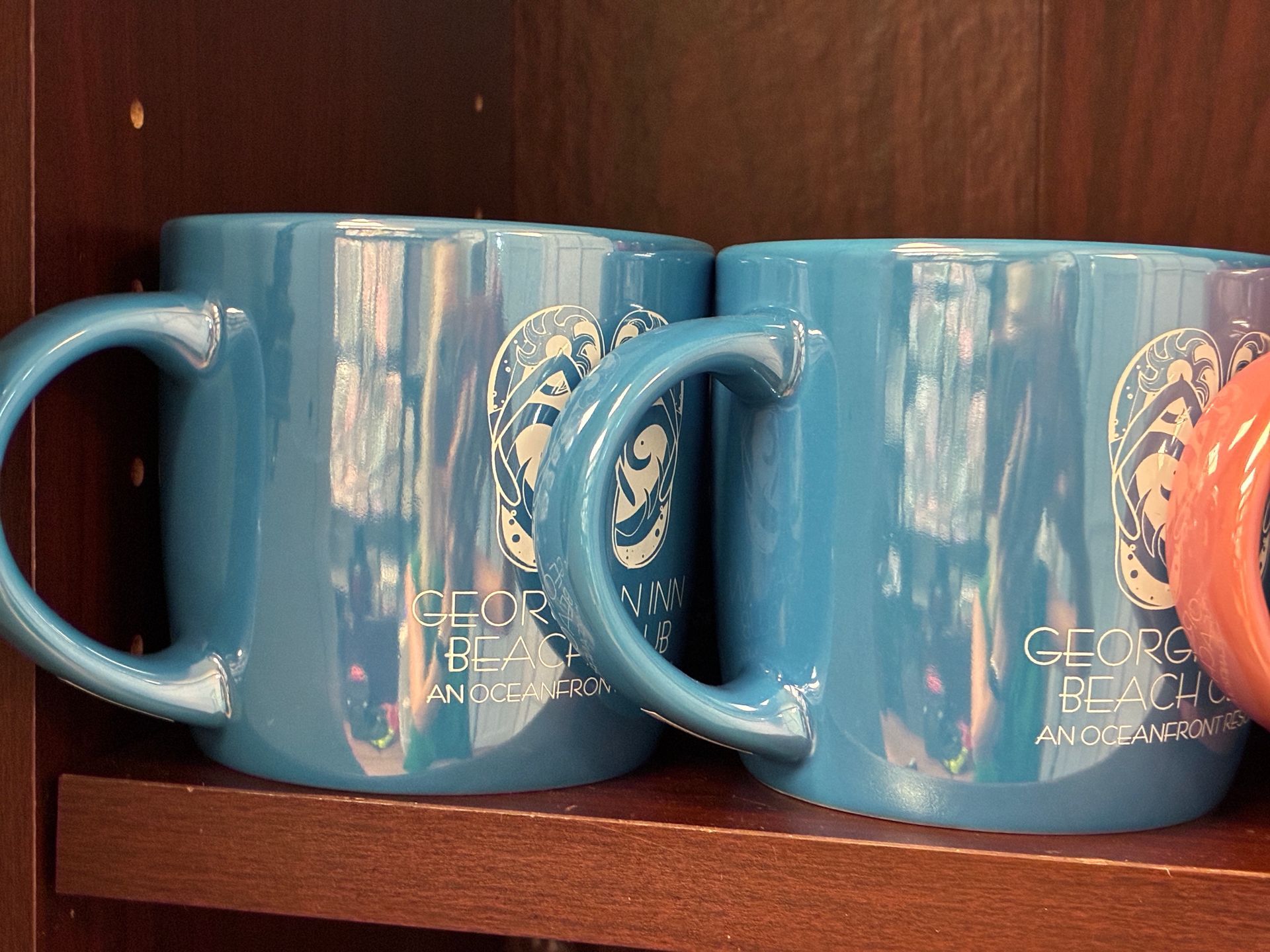 Two blue coffee mugs on a shelf with a beach-themed logo.