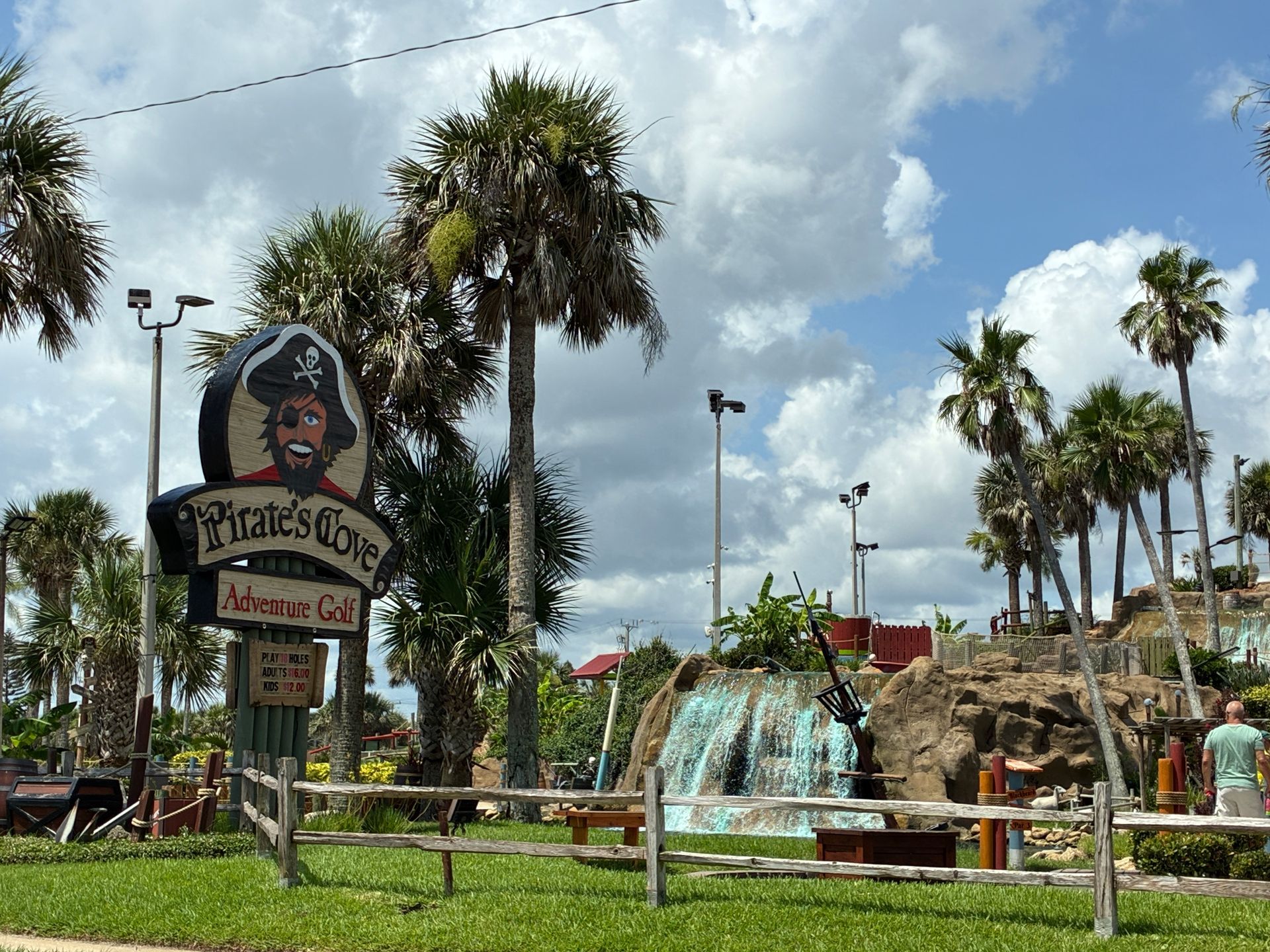 Pirate's Cove Adventure Golf sign with pirate cartoon, palm trees, waterfall, and mini golf course under blue sky.