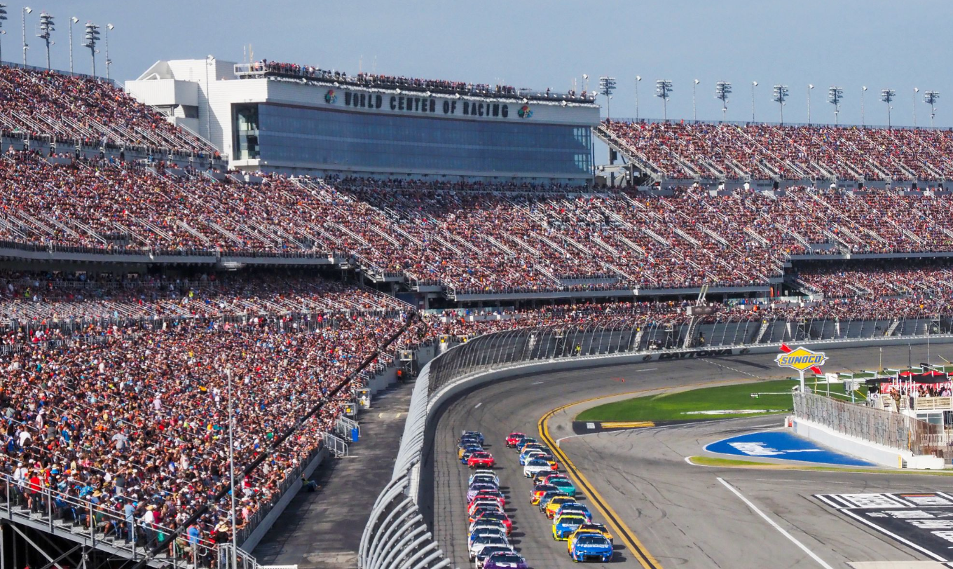 Stock car race in progress at a crowded stadium; multiple colored cars on the track.