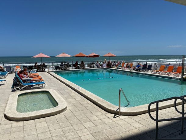 Poolside view: pool, hot tub, ocean, beach umbrellas, lounge chairs, sunny day.