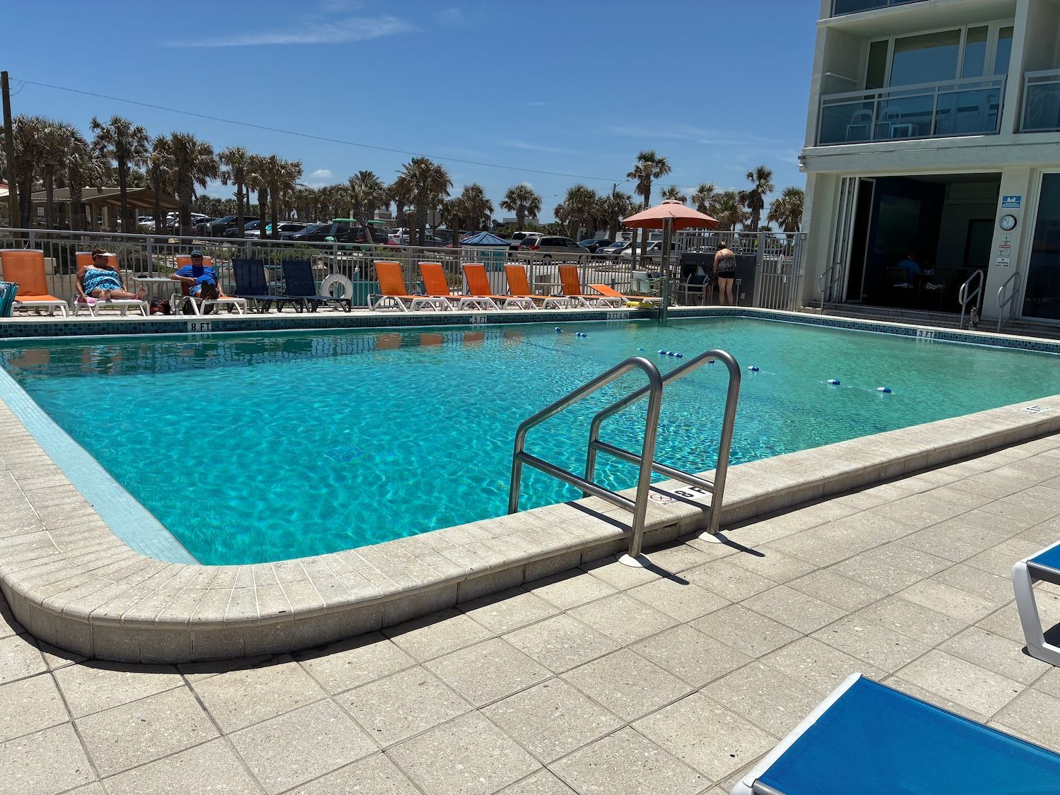 A turquoise pool with a ladder, lounge chairs, and a building, under a clear blue sky.