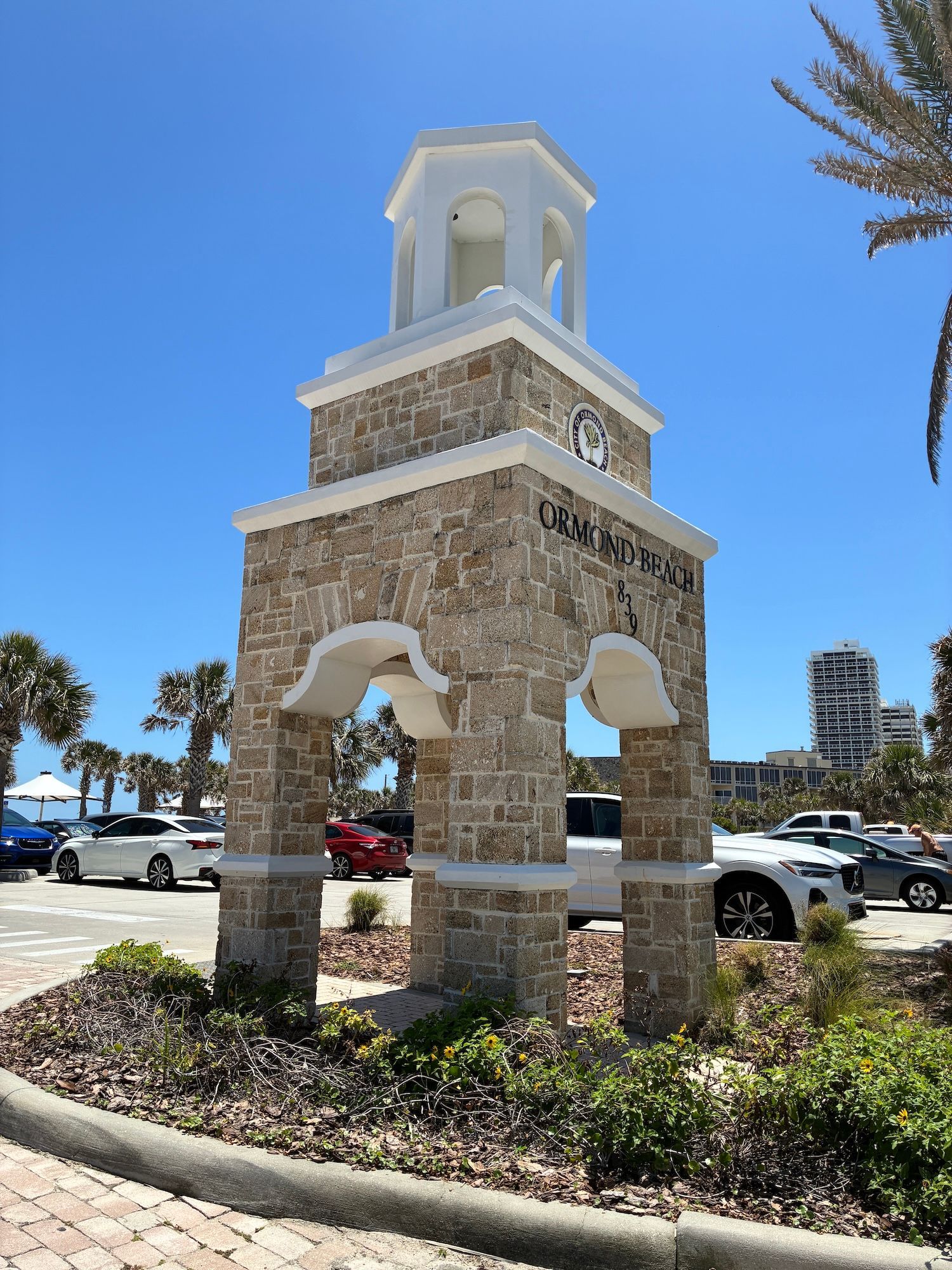 Stone tower with arched openings, clock, and white cupola under a bright blue sky; Ormond Beach sign.