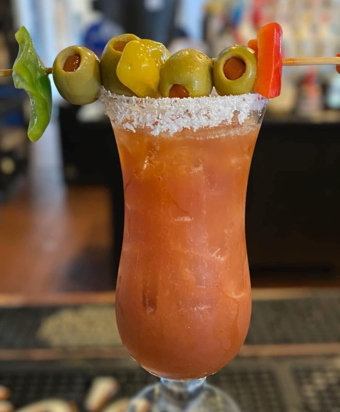 Bloody Mary cocktail in a tall glass, garnished with olives, peppers, and a salt rim.