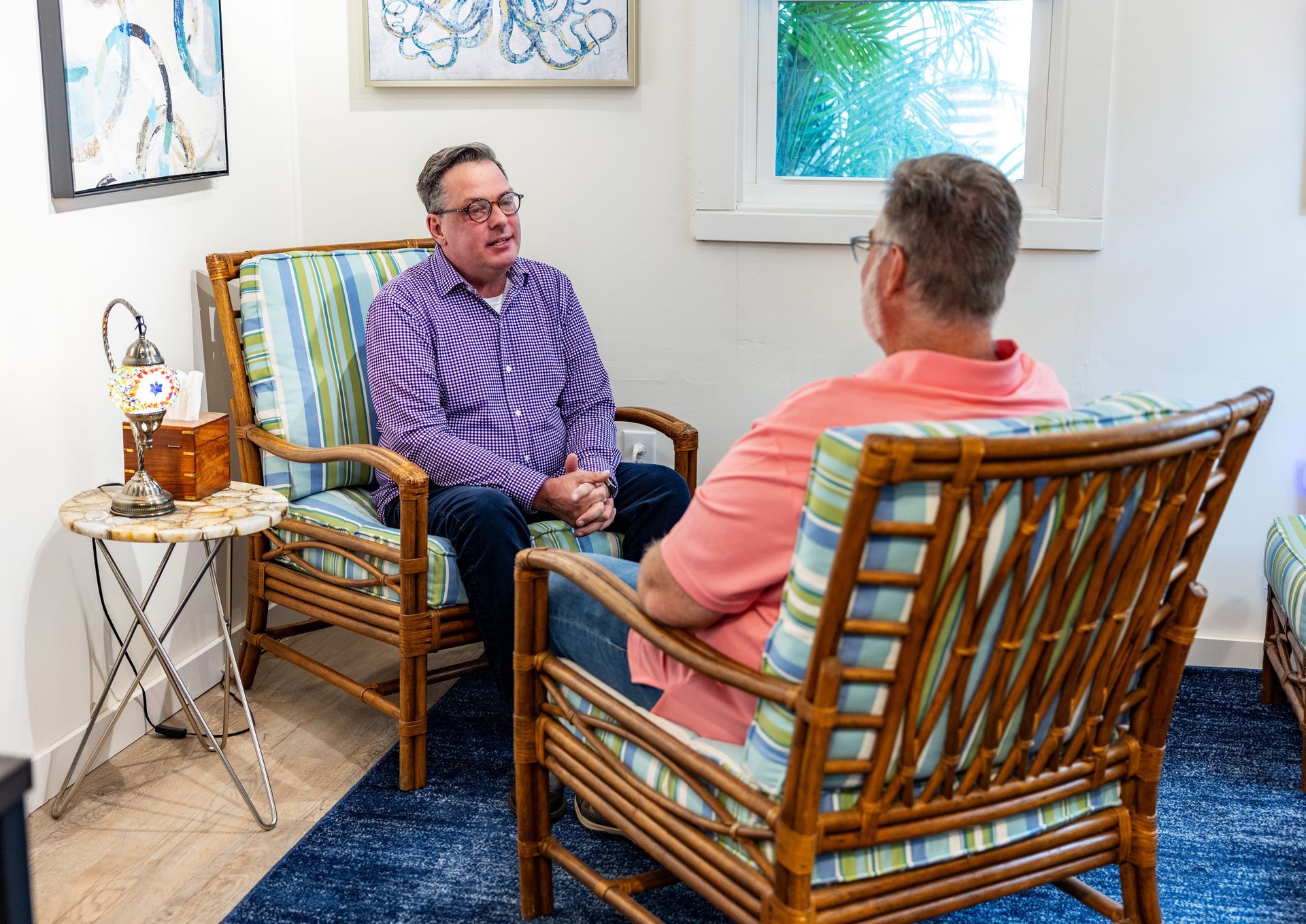 Man talking with a therapist on a sofa. Therapist holds clipboard, sunlight through window.