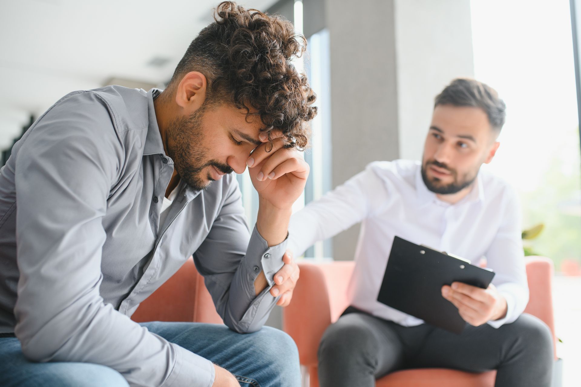 Man with hand on forehead sits with a person holding clipboard. Both are in an office setting.