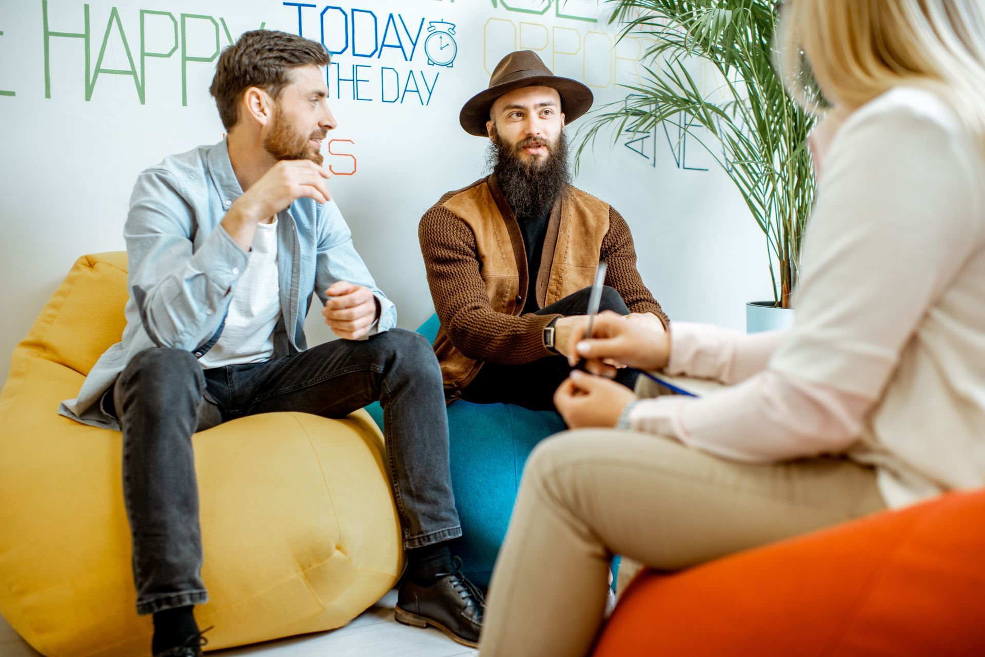 Two men in casual wear sit on beanbags, facing a woman. They are in a room with a plant and writing on the wall.
