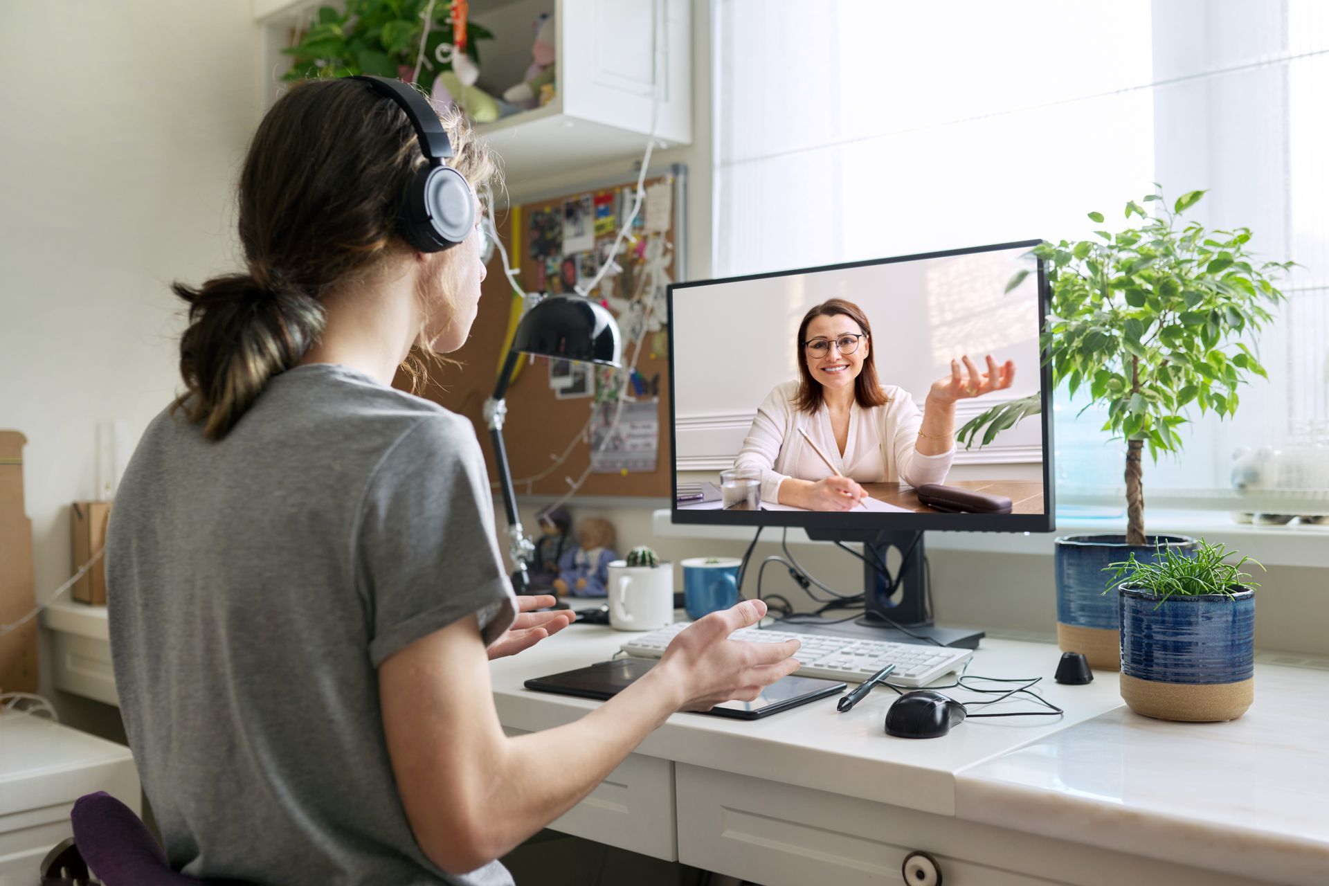Woman in headphones on a video call at a desk with a computer and plants.