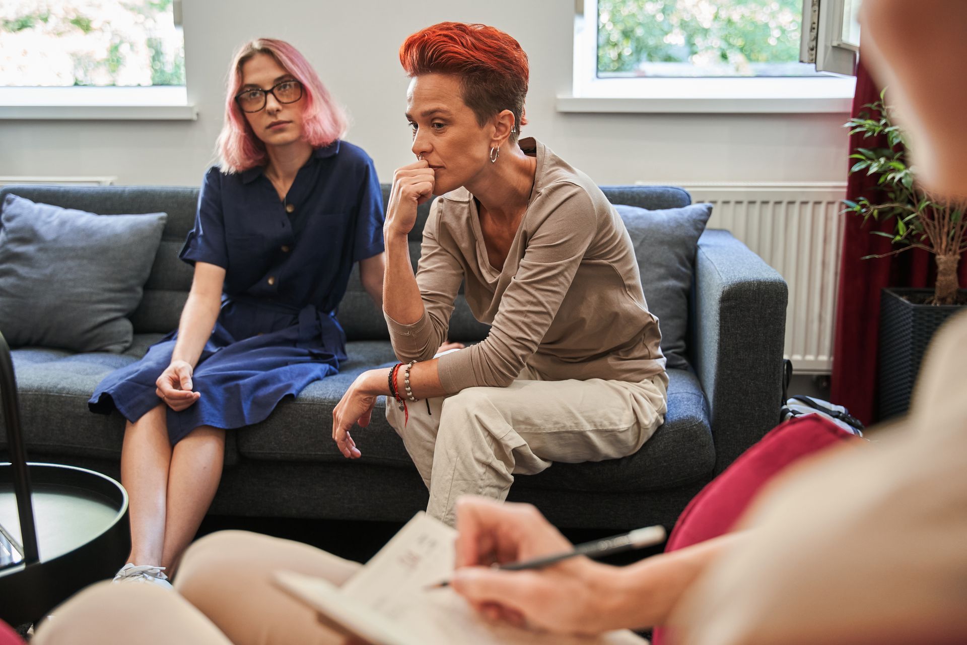 Two people on a couch look at someone taking notes. The woman on the right has red hair, and is touching her chin.