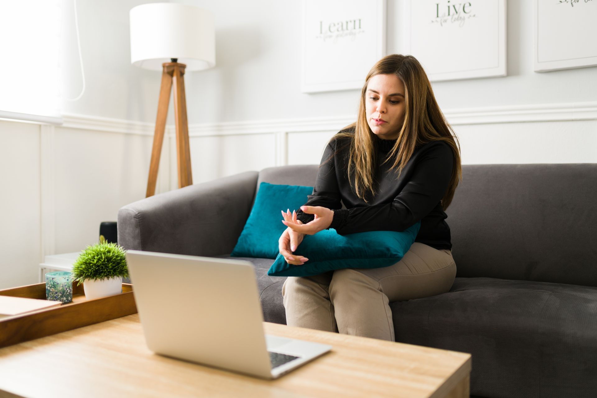 Woman on couch using a laptop, possibly in a video call. She holds a blue pillow in a living room setting.
