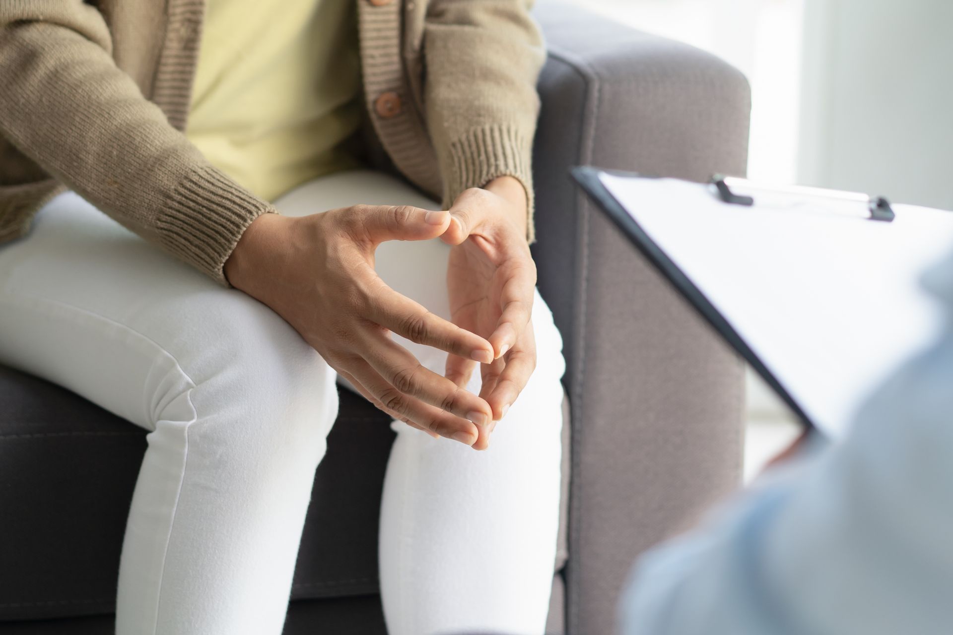 Person in neutral-colored outfit in therapy, hands clasped, facing a person holding a clipboard.