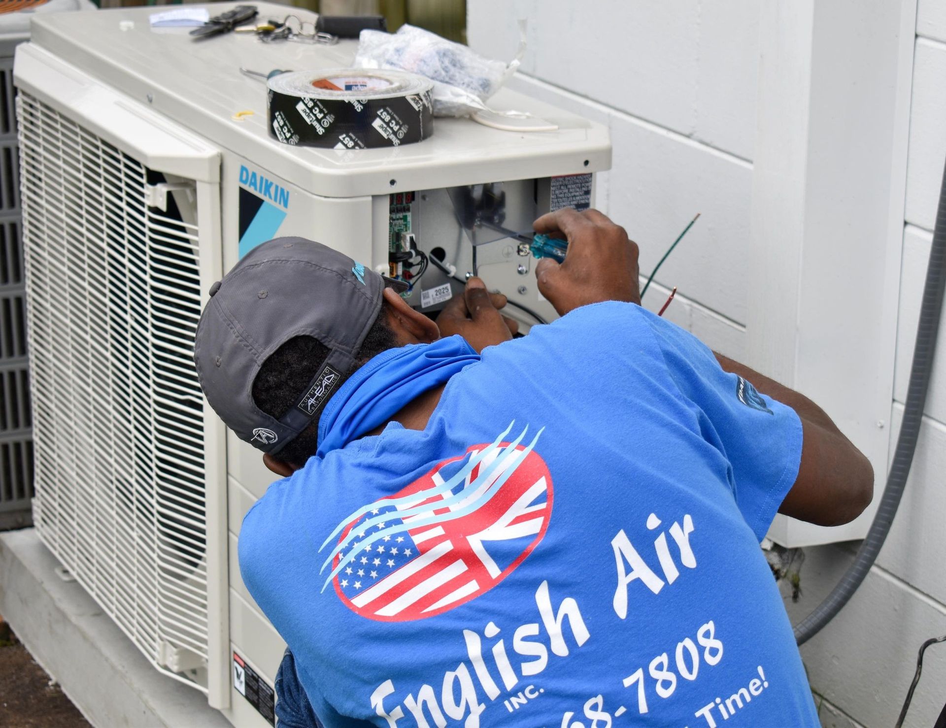 Technician in blue shirt repairing an outdoor air conditioner unit with tools nearby.