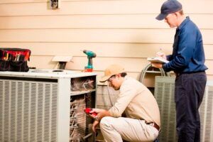 Two technicians inspect an outdoor HVAC unit beside a house, using tools and a clipboard.