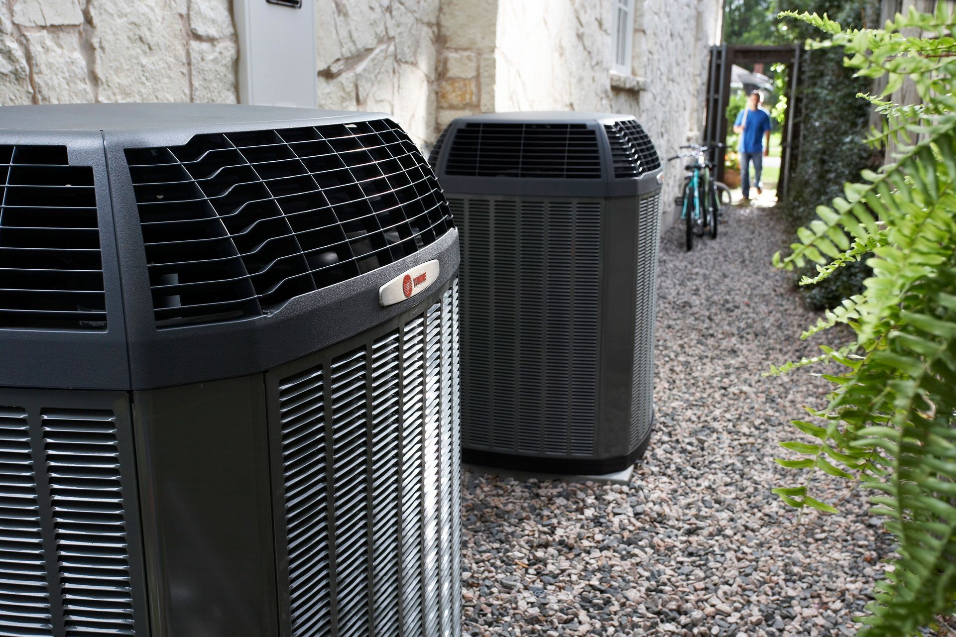 Two black outdoor HVAC units beside a stone wall along a gravel path, with ferns and a person in the distance