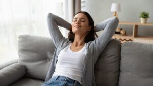 Woman relaxing on a gray couch with hands behind her head, wearing a white tank top and jeans
