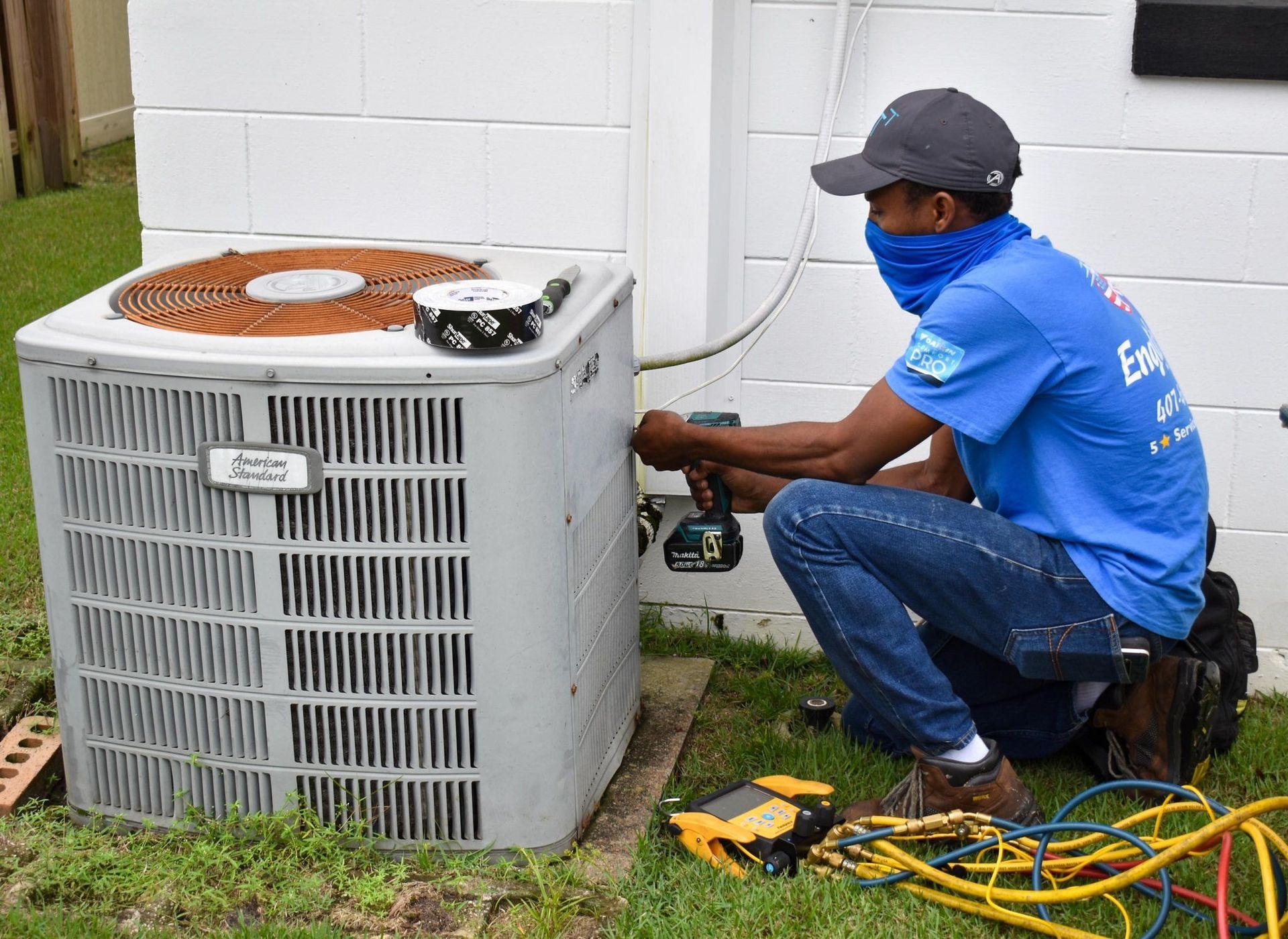 Technician in blue shirt repairing outdoor air conditioner unit with tools and hoses on grass.
