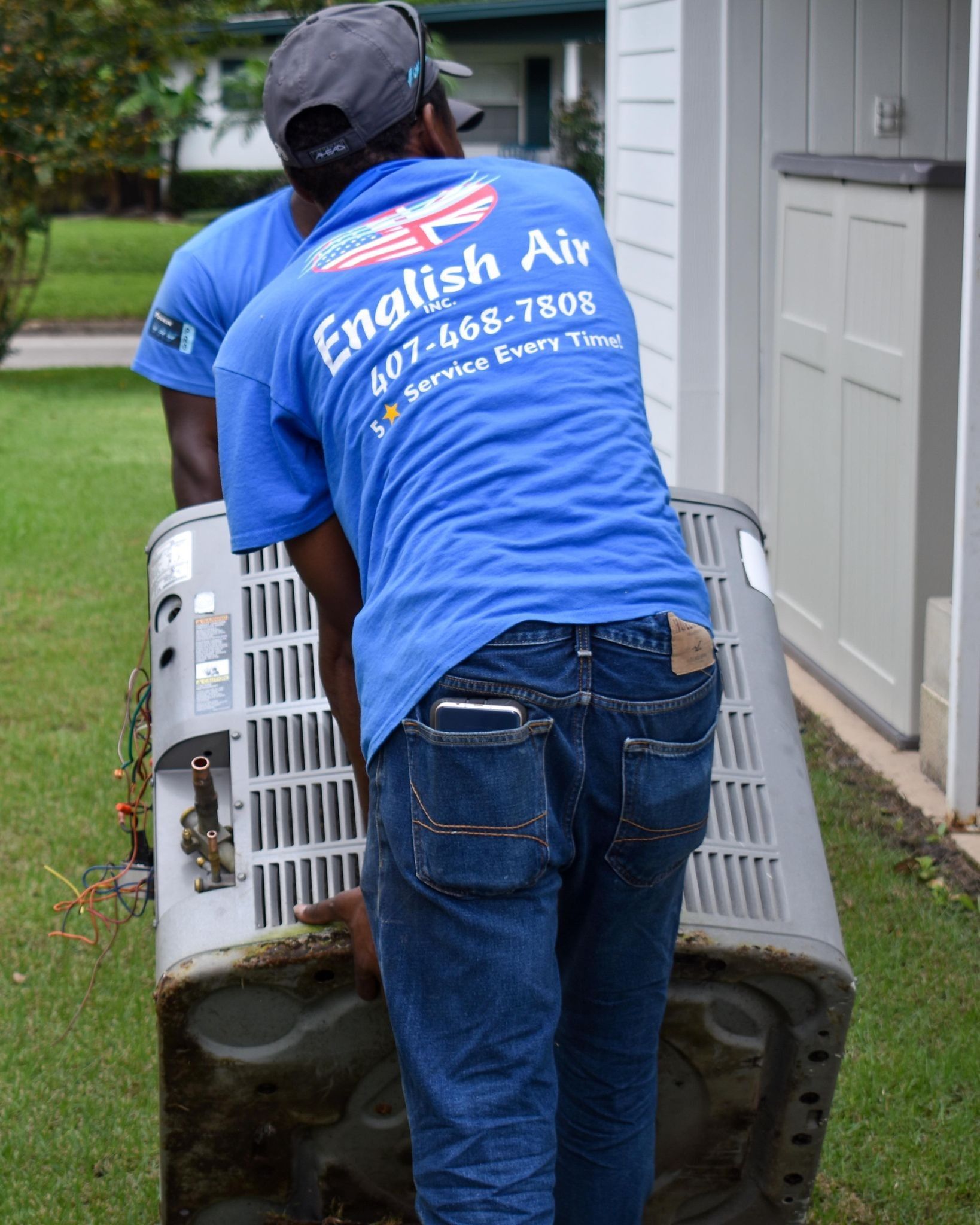 Worker in blue shirt moving a large air conditioner unit beside a house