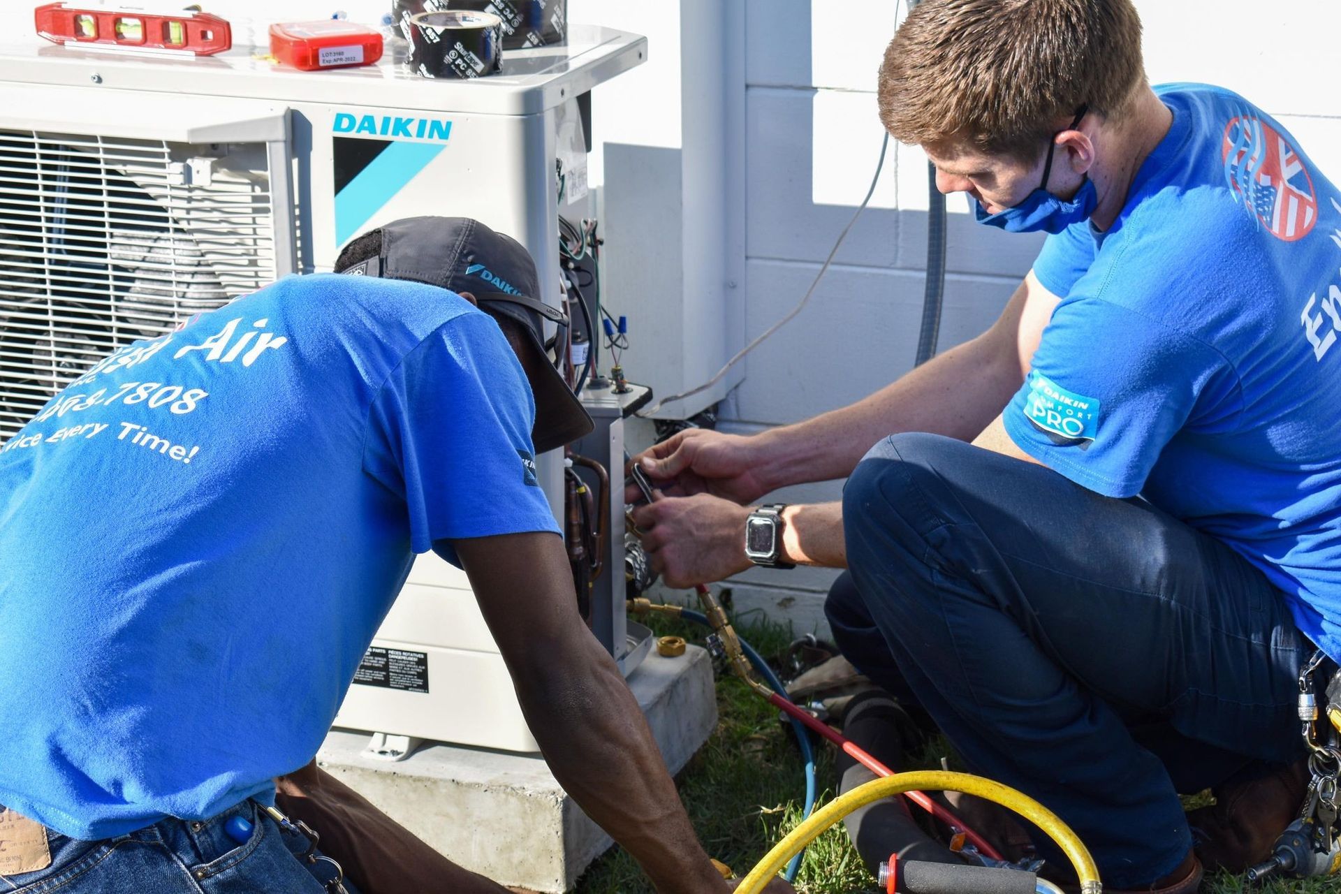 Two technicians in blue uniforms repair an outdoor air conditioner unit with tools and hoses.