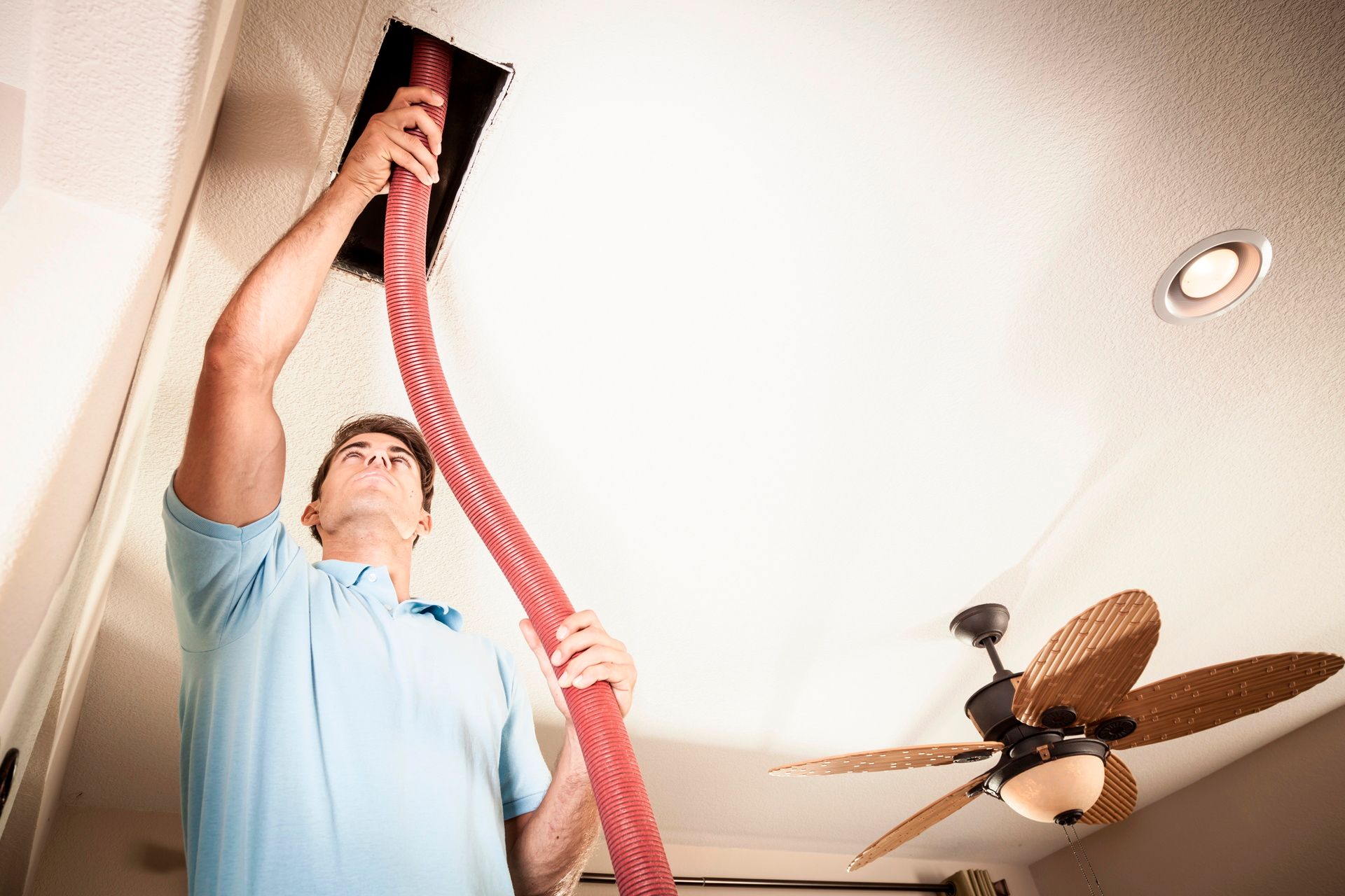 Man using a red hose to vacuum a ceiling vent in a room with a ceiling fan