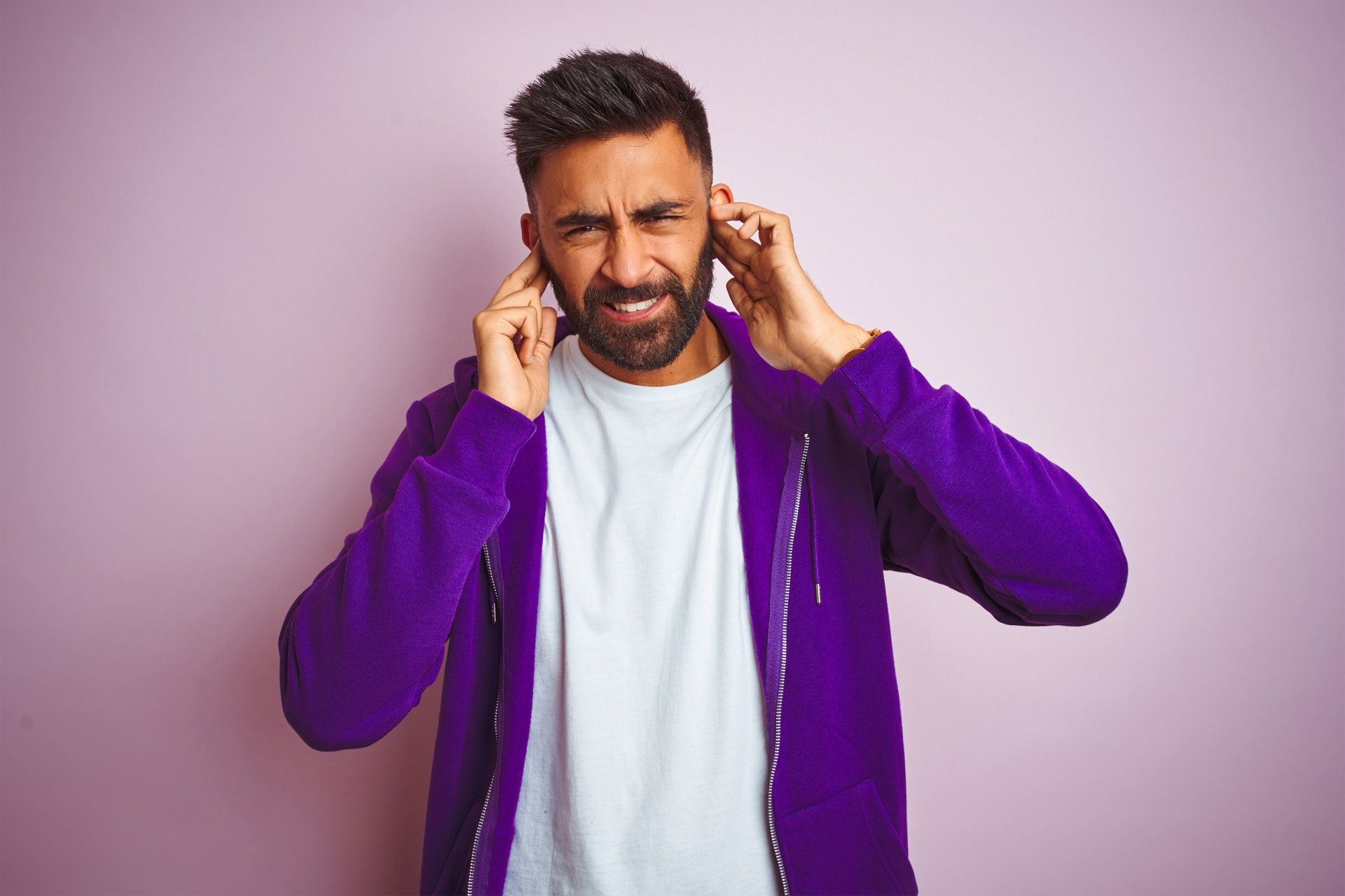 Man in a purple hoodie covering his ears, grimacing against a light purple background