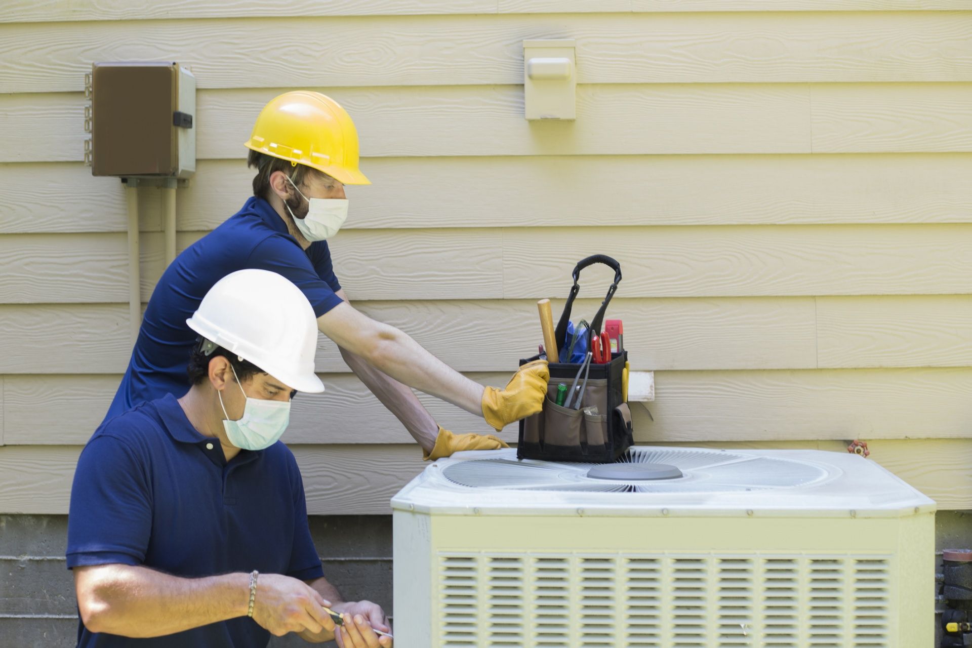Two technicians wearing hard hats service an outdoor air conditioner unit beside a house.