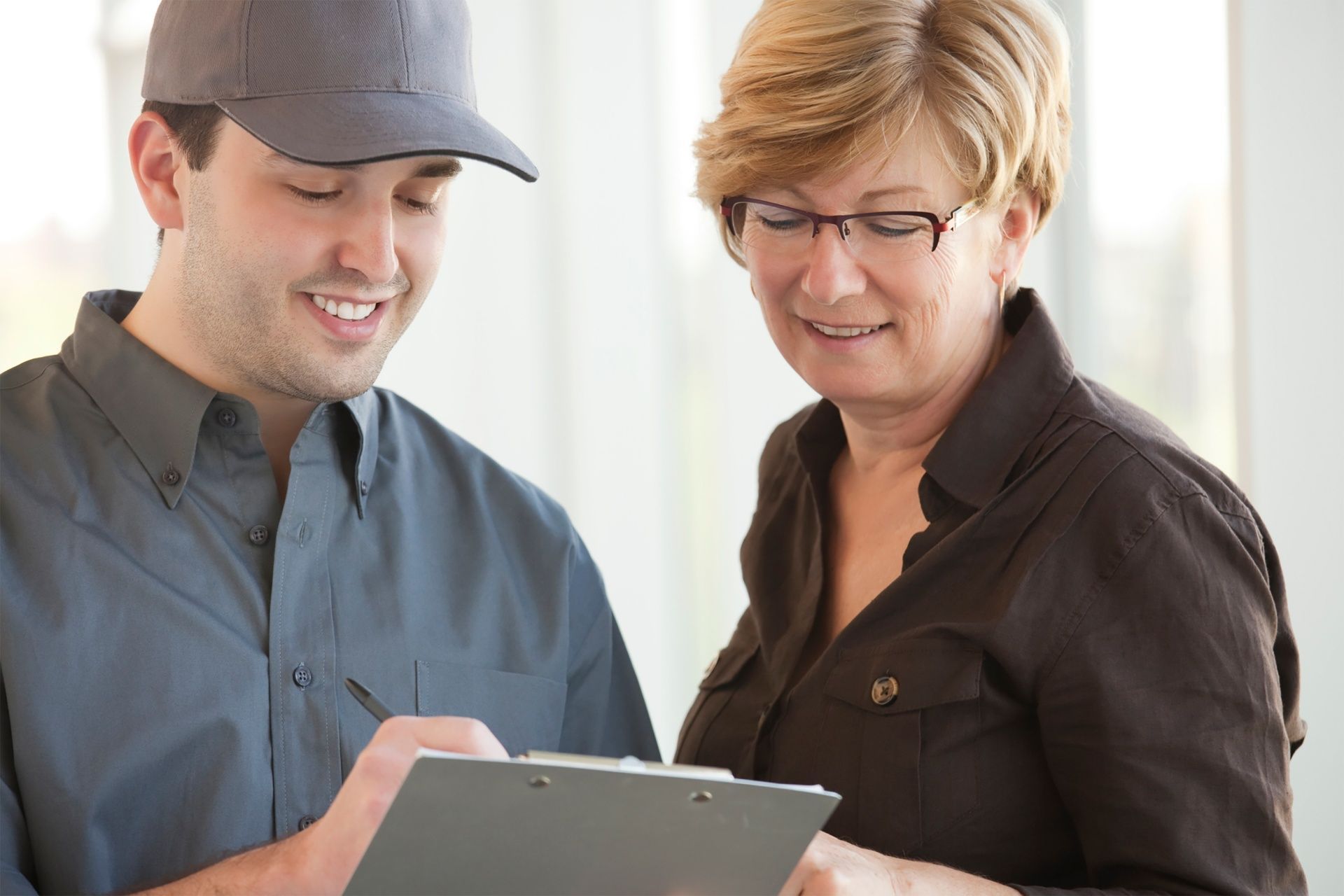 Two people reviewing information on a clipboard indoors, one in a gray cap and one in glasses and a dark shirt.