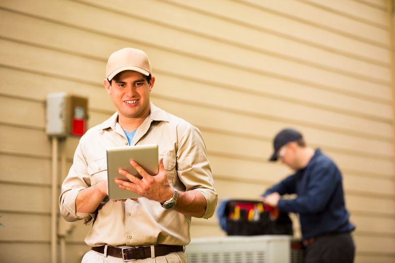 Technician holding a tablet in front of a building, with another worker using equipment in the background