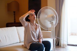 Woman sitting on a couch, wiping sweat in front of a running fan by a bright window.